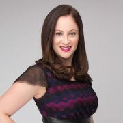 A picture of Rebecca in a purple and black dress, standing with hand on her hip, shoulder length brown hair