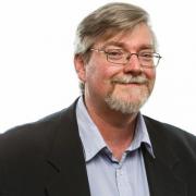 An headshot of a Keith; a man with square glasses, grey beard, and business attire